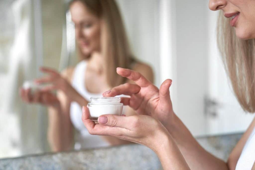 Woman dipping her finger into a face cream jar in front of a bathroom mirror, refreshing your skincare routine with a new moisturiser.