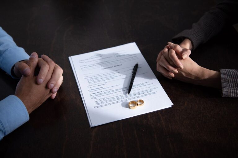 Hands of separated couple beside divorce paperwork, pen and wedding rings on a table.