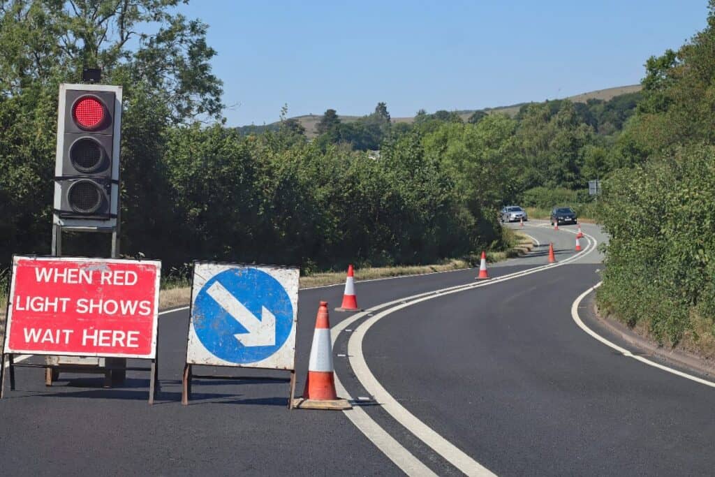 Temporary traffic lights and roadworks on a UK road with cones and a wait here sign.