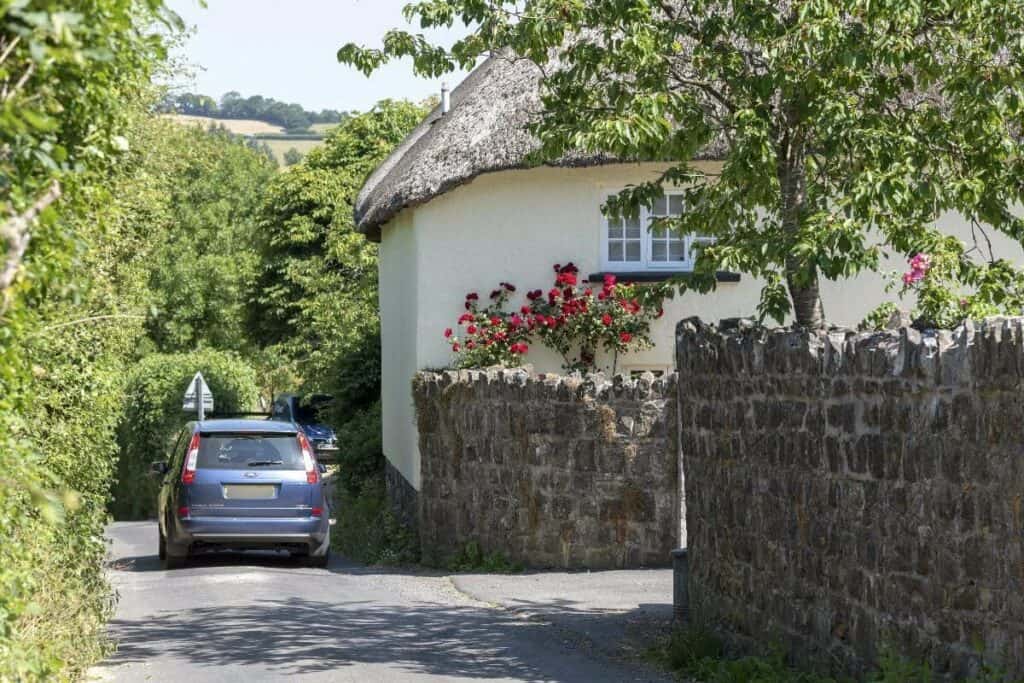 Car driving along a narrow UK country lane past a cottage, driving in the UK for the first time on rural roads