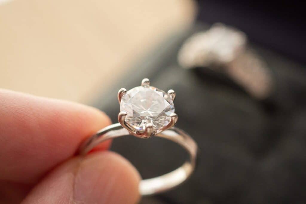 Close-up of a solitaire diamond ring held between fingers, showing the setting and prongs.