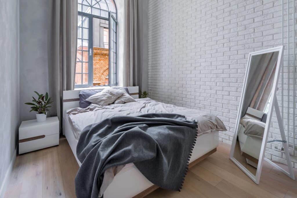Small and Bright bedroom with clear floor space, soft grey bedding, tall window and a freestanding mirror.