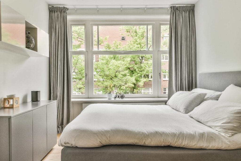 Bright bedroom with large window, soft grey curtains and minimal furniture for an open feel.