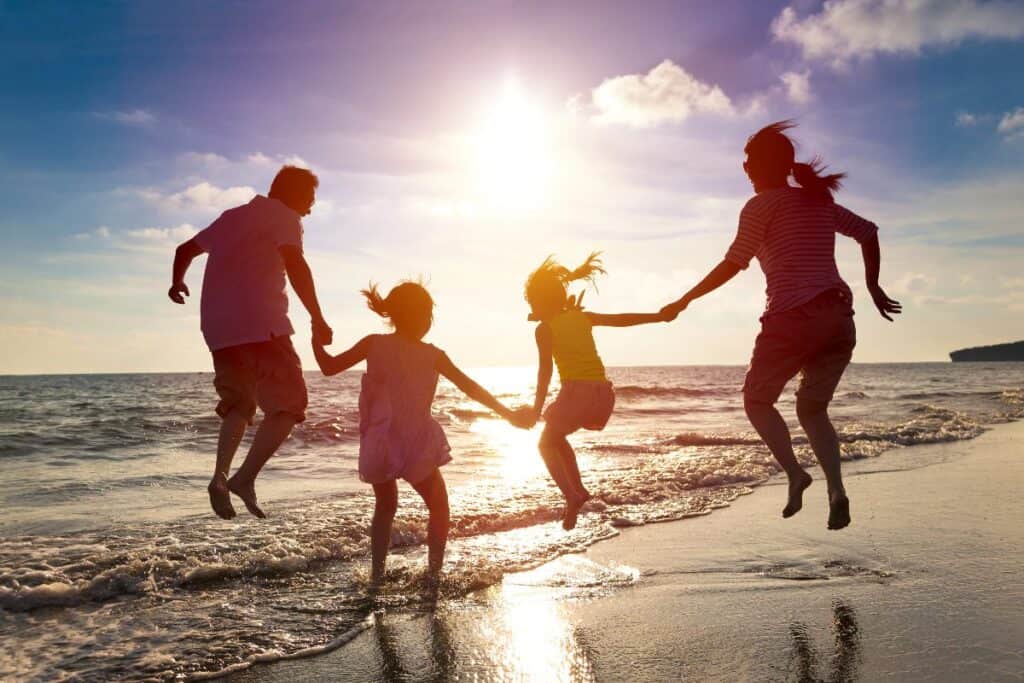 Family holding hands and jumping on a beach at sunset, showing the bonding benefits of holidays.