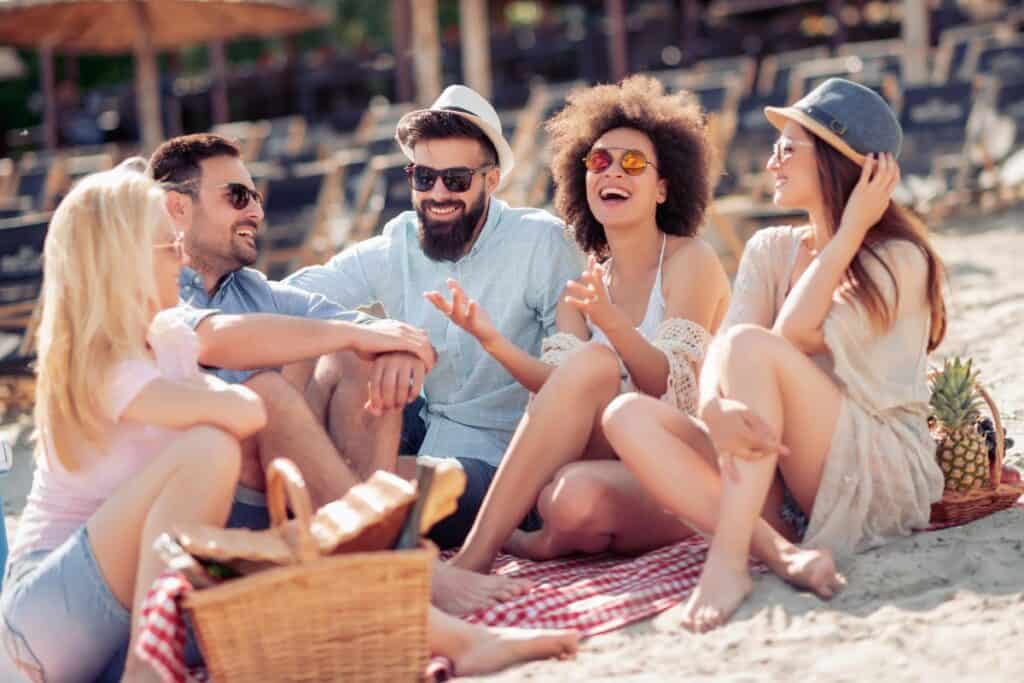 Group of friends in coordinated summer outfits laughing on a beach picnic blanket on a sunny day.