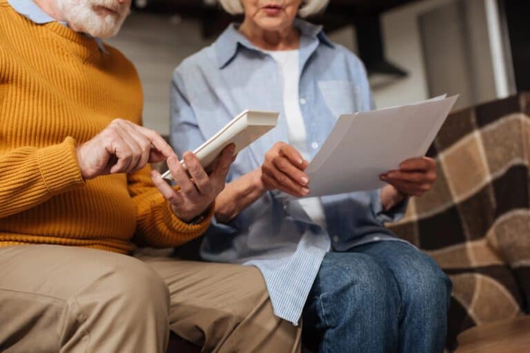 Older couple sitting together reviewing insurance paperwork and using a calculator at home.