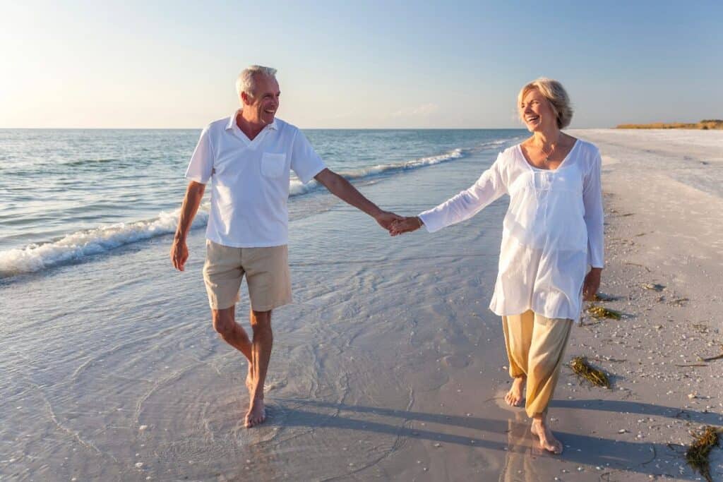 Happy older couple holding hands walking barefoot along a sunlit beach in their retirement years.