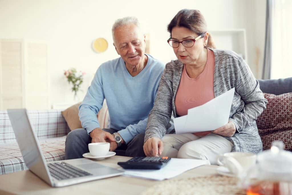 Older couple at home reviewing insurance documents on a laptop with calculator and paperwork.