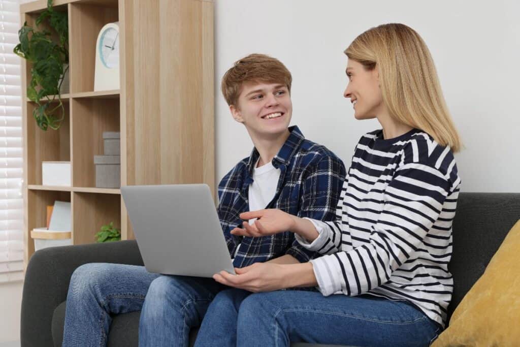 Mum and teenage son chatting on the sofa with a laptop — open communication about online safety.
