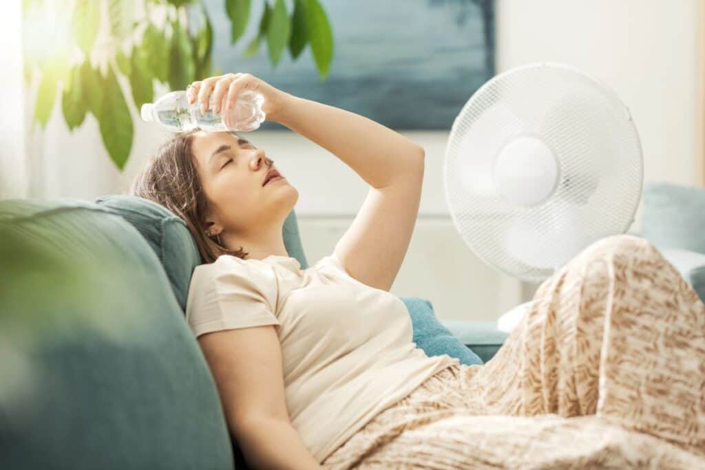 Woman cooling down with water beside a fan at home during hot weather.