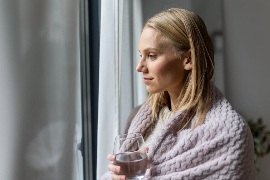 Woman resting at home by a window while planning cosmetic surgery recovery.