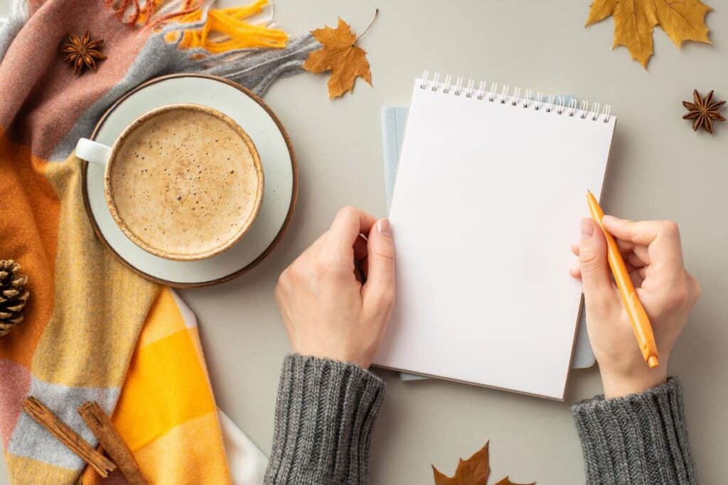 Notebook and coffee on a table with autumn leaves and cosy seasonal styling.
