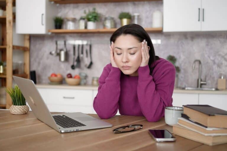 Woman sitting at a table feeling unmotivated beside a laptop and unfinished tasks.