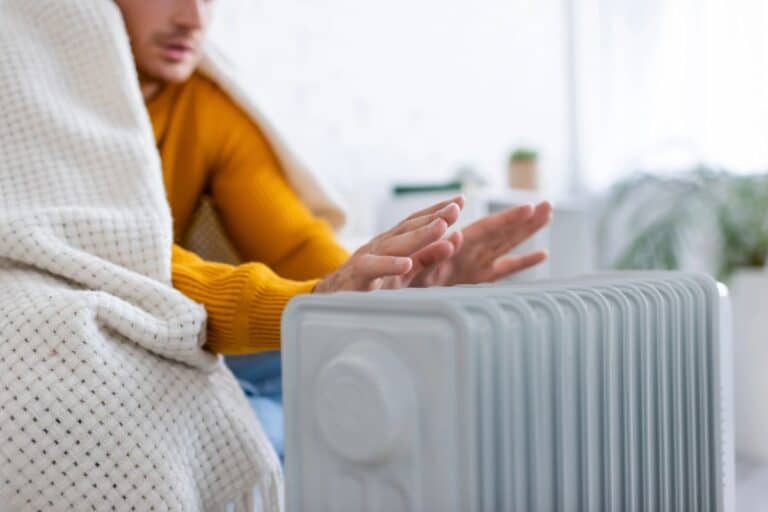 Person warming their hands by a radiator while wrapped in a blanket.