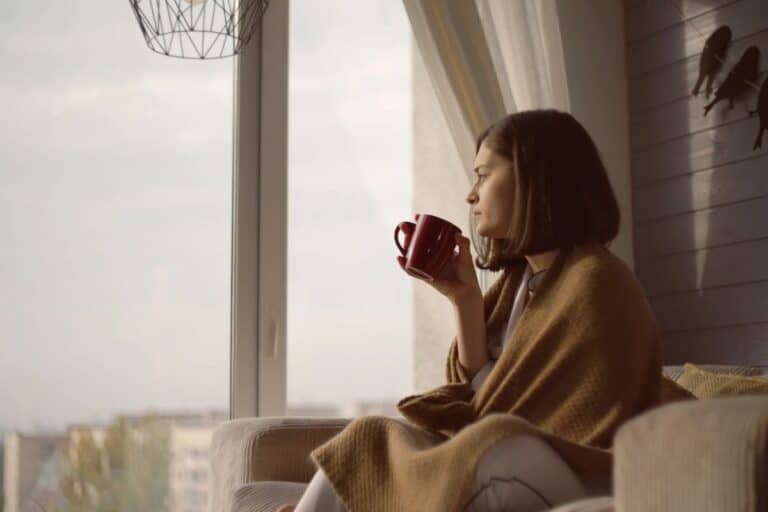 Woman wrapped in a blanket enjoying a hot drink by the window on a cosy autumn day.